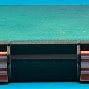 May include: A rectangular storage case featuring a dark green lid and a black base. The front of the case is secured with two metallic copper-coloured latches that contrast against the dark frame, providing a sturdy closure mechanism.