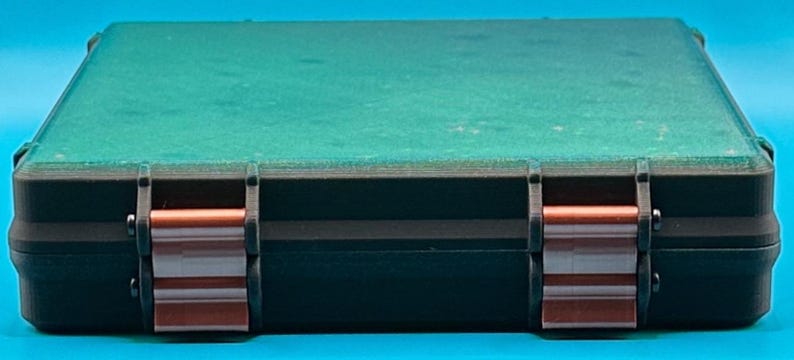 May include: A rectangular storage case featuring a dark green lid and a black base. The front of the case is secured with two metallic copper-coloured latches that contrast against the dark frame, providing a sturdy closure mechanism.