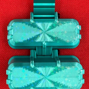 May include: A teal-colored, hinged, rectangular plastic object featuring two identical sections connected by a central pivot. Each section has a textured, iridescent surface with a star-patterned holographic finish that reflects light.