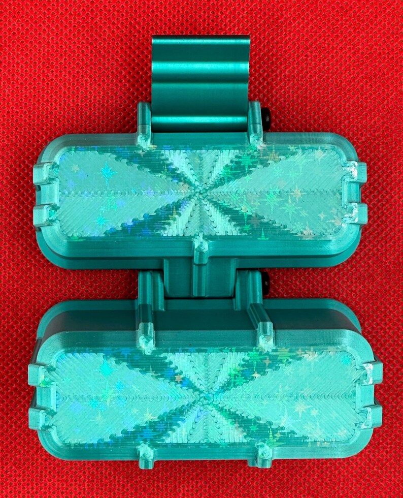 May include: A teal-colored, hinged, rectangular plastic object featuring two identical sections connected by a central pivot. Each section has a textured, iridescent surface with a star-patterned holographic finish that reflects light.