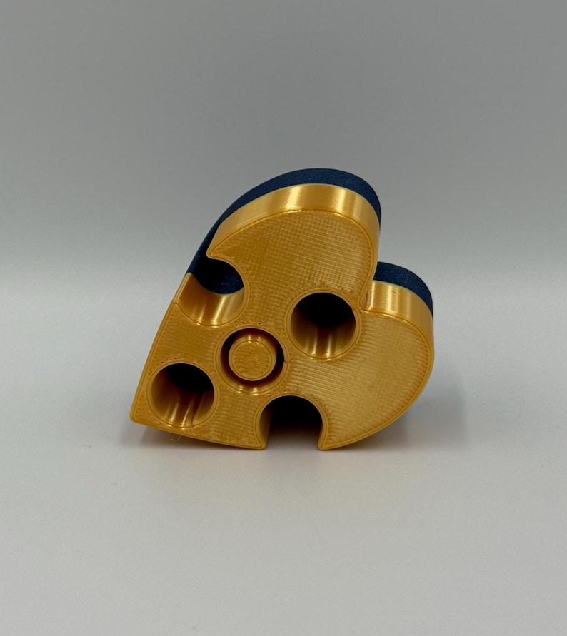 May include: A gold-colored, heart-shaped object with multiple circular openings. The top edge features a dark blue, velvety material. The object appears to be 3D-printed and has a textured surface. The design includes a central circular feature.