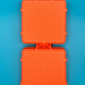 May include: A bright orange, square-shaped sandwich press featuring a hinged design with two flat, textured cooking surfaces. The appliance has a sturdy plastic exterior and a central hinge mechanism that connects the top and bottom plates.