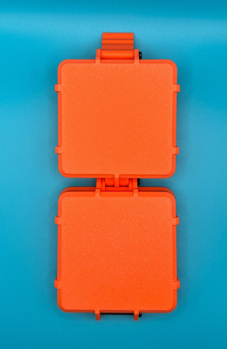 May include: A bright orange, square-shaped sandwich press featuring a hinged design with two flat, textured cooking surfaces. The appliance has a sturdy plastic exterior and a central hinge mechanism that connects the top and bottom plates.