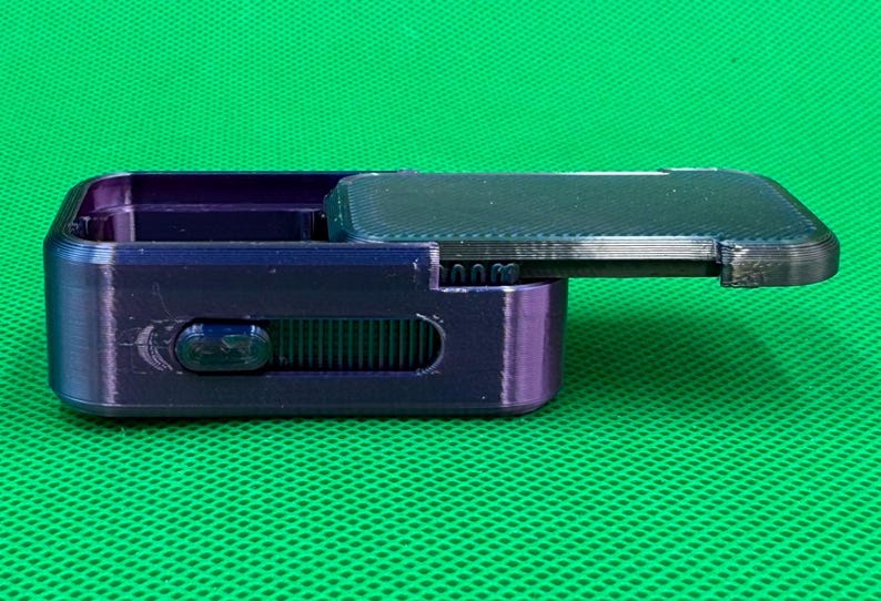 May include: A dark purple, rectangular box with a hinged lid, open to reveal the interior. The box has a sliding mechanism on the side and is set against a green background. The lid is textured.