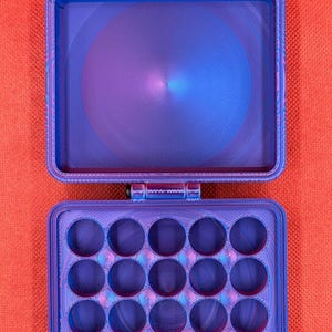 May include: A rectangular, hinged storage case with a vibrant blue and purple gradient finish. The top section is an open, empty compartment, while the bottom section features a grid of sixteen circular cutouts for organising small items.