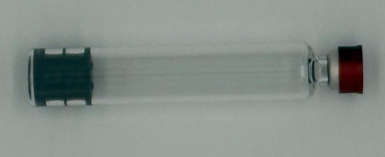 May include: A clear glass cylindrical vial featuring a dark green rubber stopper at one end and a metallic silver neck topped with a small, flat, circular red cap at the opposite end. The vial is empty and has a smooth, transparent body.