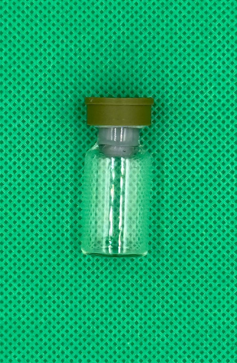 May include: A small, clear glass vial featuring a cylindrical body and a flat base. The vial is topped with a grey rubber stopper and a solid olive-green metal cap, creating a secure seal for the container's opening at the top.