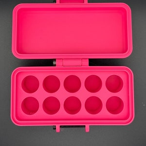 May include: A bright pink rectangular storage case with a hinged lid. The interior base features ten circular recessed compartments arranged in two rows of five, designed for holding small items securely within the open container.