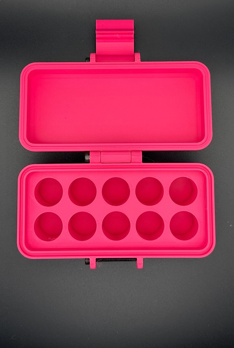 May include: A bright pink rectangular storage case with a hinged lid. The interior base features ten circular recessed compartments arranged in two rows of five, designed for holding small items securely within the open container.