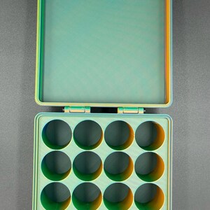 May include: A green and orange plastic storage box with a hinged lid and 15 round compartments. The box is open and the lid is propped up.