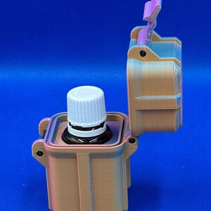 May include: A tan, rectangular, 3D-printed storage case with a hinged lid standing open. Inside the case sits a small, dark amber glass bottle with a white, ribbed, screw-top cap. The case features vertical ridges and a latch mechanism.