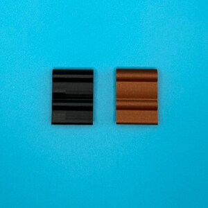 May include: Two rectangular clips with a curved, wave-like profile. One clip is solid black and the other is a metallic copper color. Both items feature a series of horizontal ridges along their surface, providing a textured appearance.