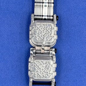 K&ouml;nnte beinhalten: Ein silberfarbenes Metall-Uhrenarmband mit zwei quadratischen, texturierten Gliedern in einem sich wiederholenden geometrischen Muster. Das Band enth&auml;lt einen gestreiften, rechteckigen oberen Anschluss und einen schwarzen Schraubstift.