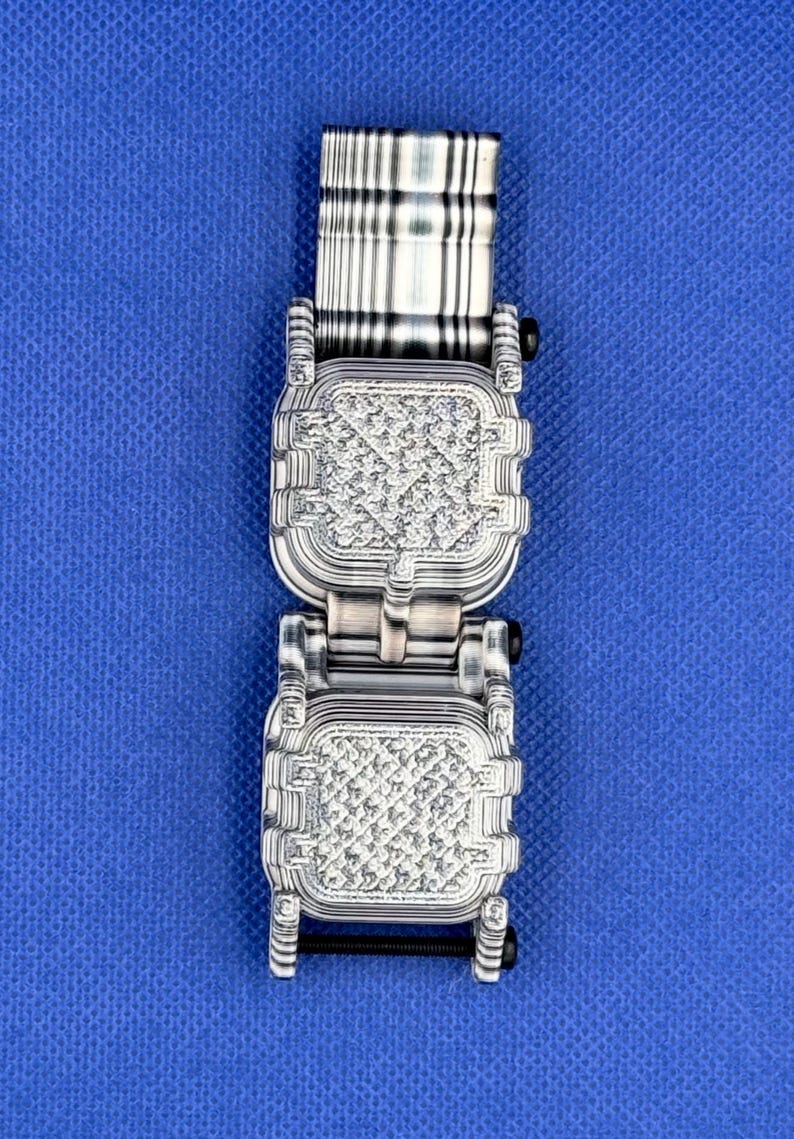 K&ouml;nnte beinhalten: Ein silberfarbenes Metall-Uhrenarmband mit zwei quadratischen, texturierten Gliedern in einem sich wiederholenden geometrischen Muster. Das Band enth&auml;lt einen gestreiften, rechteckigen oberen Anschluss und einen schwarzen Schraubstift.