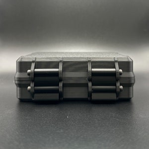May include: A rectangular black storage case with a textured top surface and two prominent hinged locking latches on the front side. The case features a layered, matte finish with visible horizontal lines consistent with 3D printing.