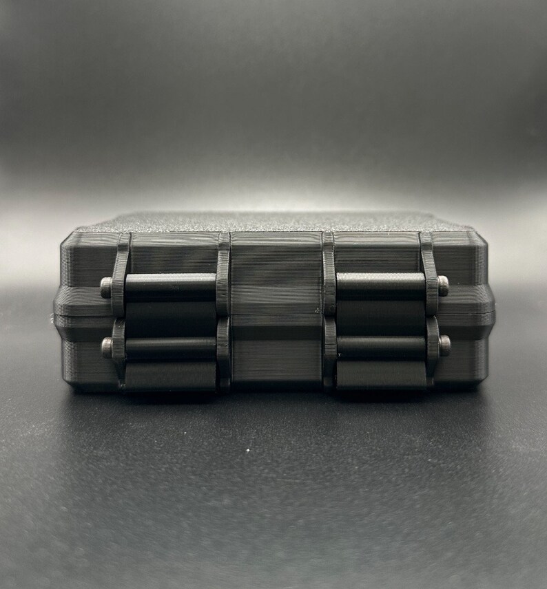May include: A rectangular black storage case with a textured top surface and two prominent hinged locking latches on the front side. The case features a layered, matte finish with visible horizontal lines consistent with 3D printing.