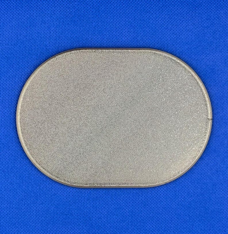 May include: A silver-colored oval patch with a textured, glittery surface and a stitched border. The object features a uniform metallic finish across its entire front face, presenting a clean, simple shape with rounded edges and a flat profile.