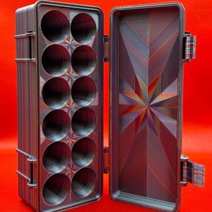 May include: A rectangular storage case with a hinged lid, featuring a multi-coloured striped pattern. The interior contains twelve circular compartments arranged in two columns, while the inner lid displays a geometric starburst design.