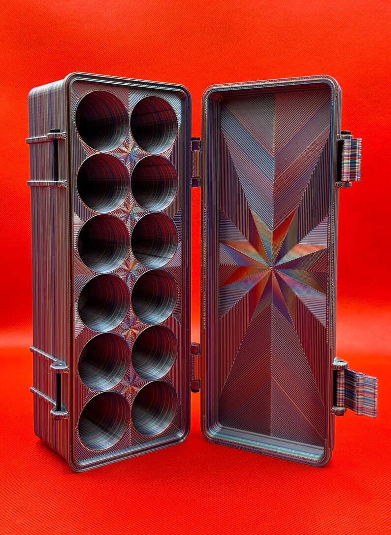 May include: A rectangular storage case with a hinged lid, featuring a multi-coloured striped pattern. The interior contains twelve circular compartments arranged in two columns, while the inner lid displays a geometric starburst design.