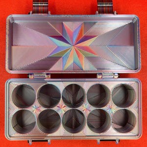 Puede incluir: Un estuche de almacenamiento rectangular de metal iridiscente con tapa abatible. El interior presenta un patr&oacute;n de estrella en la tapa y diez compartimentos circulares dispuestos en dos filas de cinco, con un acabado lineal multicolor.