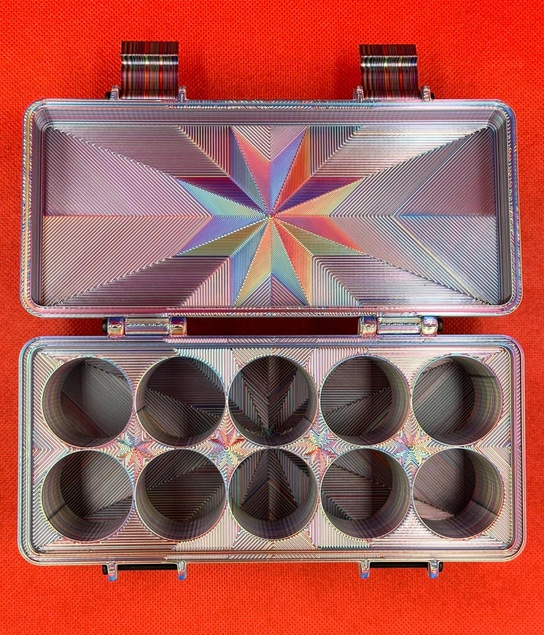 Puede incluir: Un estuche de almacenamiento rectangular de metal iridiscente con tapa abatible. El interior presenta un patr&oacute;n de estrella en la tapa y diez compartimentos circulares dispuestos en dos filas de cinco, con un acabado lineal multicolor.