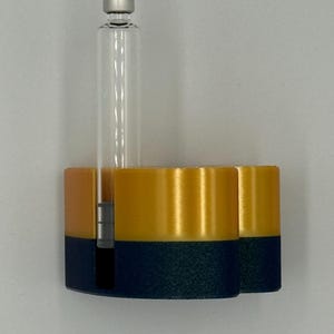 May include: A 3D-printed, two-tone holder in navy blue and gold, with a clear glass vial and silver cap. The holder has two cylindrical compartments and a slot for the vial. The design is modern and minimalist.
