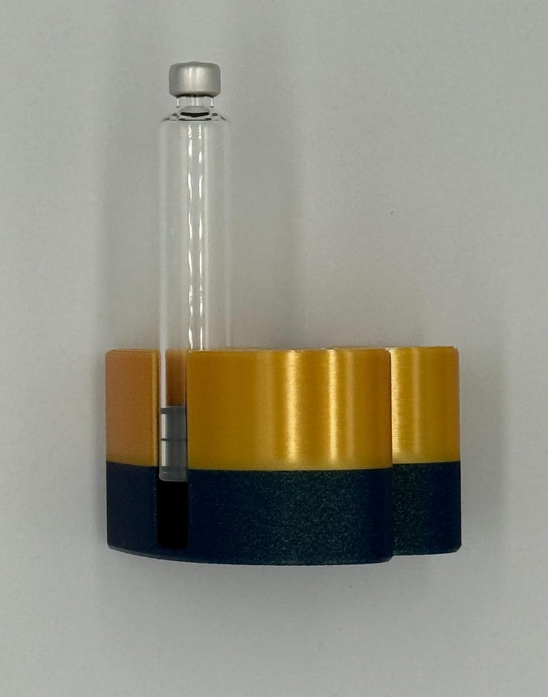May include: A 3D-printed, two-tone holder in navy blue and gold, with a clear glass vial and silver cap. The holder has two cylindrical compartments and a slot for the vial. The design is modern and minimalist.