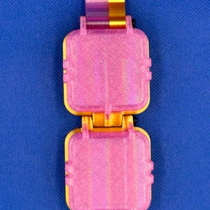 May include: A hinged, rectangular container featuring two square, glittery pink sections connected by a central yellow and pink mechanism. The object has a textured, layered surface finish and small black hardware components at the joints.