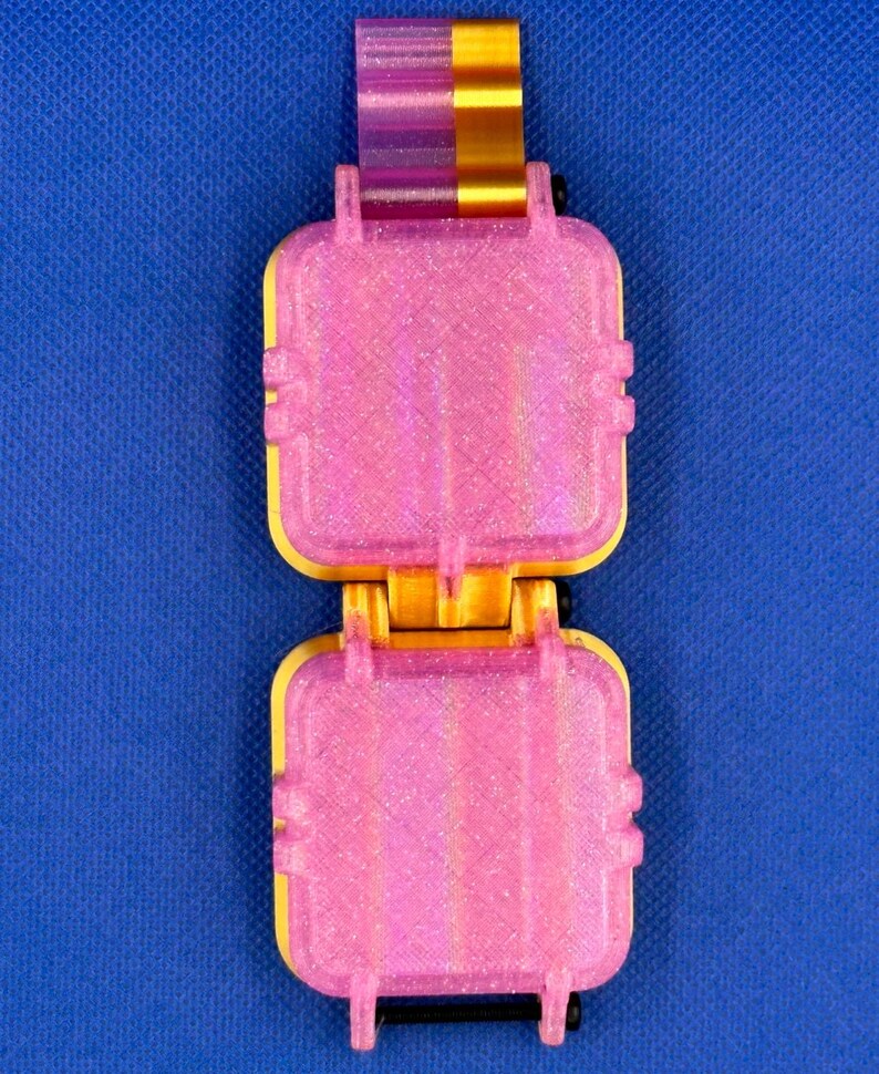 May include: A hinged, rectangular container featuring two square, glittery pink sections connected by a central yellow and pink mechanism. The object has a textured, layered surface finish and small black hardware components at the joints.