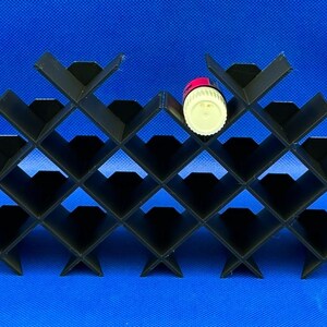 Puede incluir: Un botellero negro modular con patr&oacute;n de rombos y m&uacute;ltiples compartimentos. Una peque&ntilde;a botella cil&iacute;ndrica con tap&oacute;n blanco y etiqueta roja est&aacute; colocada en uno de los compartimentos centrales superiores del estante.