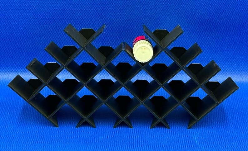 Puede incluir: Un botellero negro modular con patr&oacute;n de rombos y m&uacute;ltiples compartimentos. Una peque&ntilde;a botella cil&iacute;ndrica con tap&oacute;n blanco y etiqueta roja est&aacute; colocada en uno de los compartimentos centrales superiores del estante.