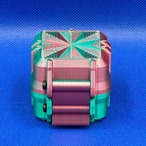 May include: A small, square, 3D-printed box featuring a gradient of teal and magenta colors. The lid displays a geometric starburst pattern with layered ridges, while the sides have a smooth, vertical texture and two small black circular fasteners.