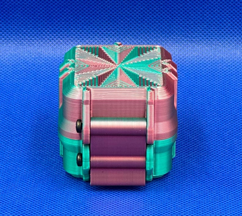 May include: A small, square, 3D-printed box featuring a gradient of teal and magenta colors. The lid displays a geometric starburst pattern with layered ridges, while the sides have a smooth, vertical texture and two small black circular fasteners.
