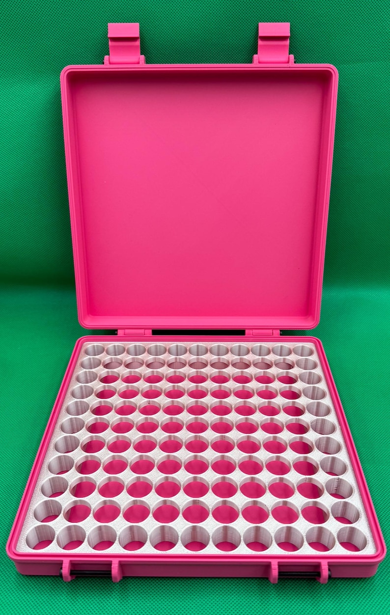 K&ouml;nnte beinhalten: Ein leuchtend pinkfarbener Aufbewahrungskoffer aus Kunststoff mit offenem Deckel und einem herausnehmbaren Inneneinsatz, der ein Raster aus einhundert kreisf&ouml;rmigen L&ouml;chern aufweist. Der Koffer hat ein Klappdesign mit zwei Verschl&uuml;ssen.