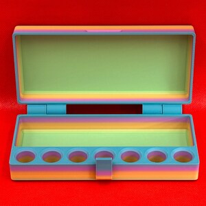 May include: A rectangular, open storage box featuring a gradient color scheme that transitions from orange and pink to blue. The interior base includes seven circular cutouts for holding small items, while the lid remains open.