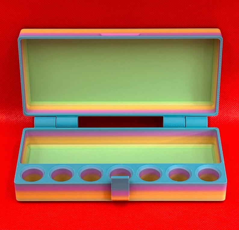 May include: A rectangular, open storage box featuring a gradient color scheme that transitions from orange and pink to blue. The interior base includes seven circular cutouts for holding small items, while the lid remains open.