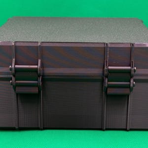 May include: A rectangular storage case with a dark, multi-toned purple and black finish. The box features a textured lid and two prominent vertical latches on the front panel, designed with a rigid, structured exterior and a flat top surface.