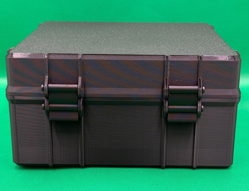 May include: A rectangular storage case with a dark, multi-toned purple and black finish. The box features a textured lid and two prominent vertical latches on the front panel, designed with a rigid, structured exterior and a flat top surface.