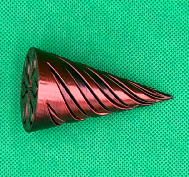 May include: A metallic red, conical object with a spiraling, grooved texture along its length. The flat, circular base features a recessed, symmetrical four-petal flower design. The object tapers to a pointed tip, showcasing a smooth finish.