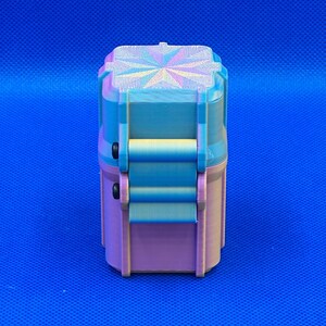 May include: A small, rectangular, 3D-printed storage container with a hinged lid. The object features a gradient color scheme transitioning from light blue at the top to a soft pink at the base, with a geometric star pattern on the lid.