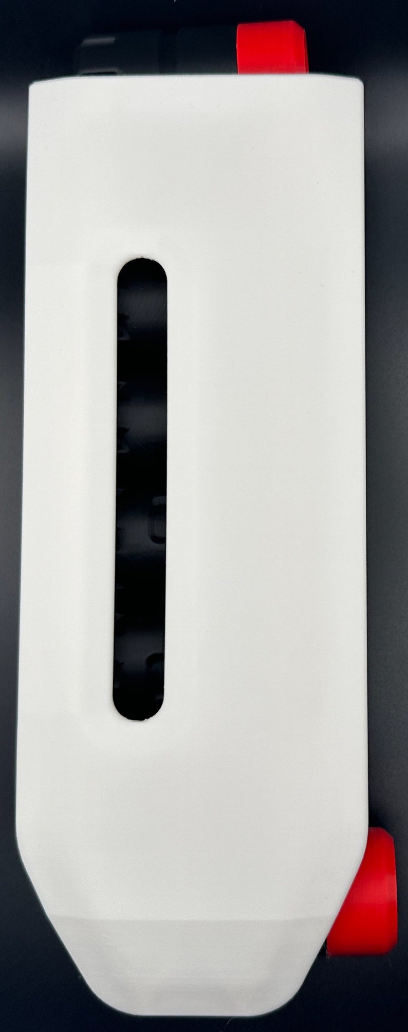 May include: A white rectangular dispenser featuring a vertical black viewing slot in the center. The unit includes a bright red cap at the top and a matching red circular component protruding from the bottom right side.