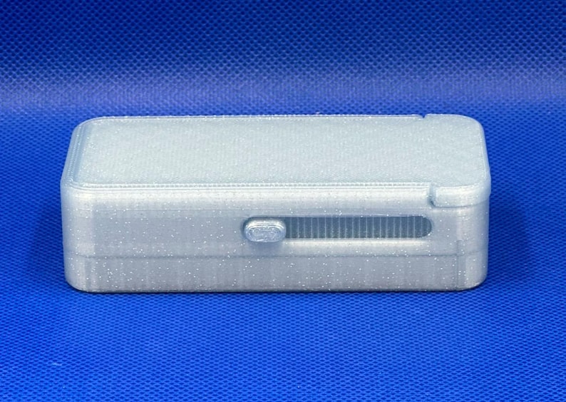 May include: A rectangular, silver-colored storage box with a glittery finish. The box features a sliding latch mechanism on the front side, which is positioned within a horizontal recessed track. The lid is flat and the edges are rounded.