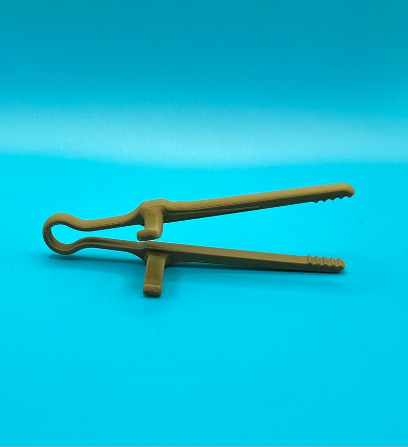 May include: A brown plastic kitchen tong tool featuring a looped handle at one end and two long, straight arms with serrated, gripping tips. The design includes a small, perpendicular support tab on each arm to keep the tool elevated.