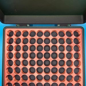 May include: A black storage case with a hinged lid featuring the word Glow in metallic orange script. The interior contains a grid of 81 circular openings with orange walls and black bases, designed for organising small items in a square layout.