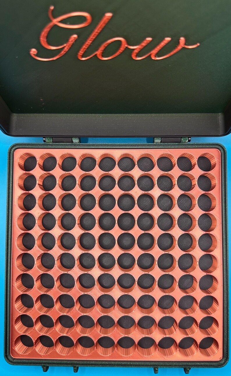 May include: A black storage case with a hinged lid featuring the word Glow in metallic orange script. The interior contains a grid of 81 circular openings with orange walls and black bases, designed for organising small items in a square layout.
