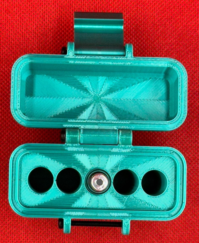 May include: A teal 3D-printed rectangular storage case with a hinged lid. The interior base features four circular compartments and one central cylindrical slot containing a small metal component with a circular opening.