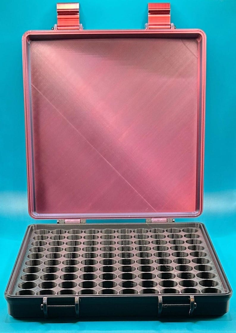 May include: A rectangular storage case with a metallic red lid and a black base. The interior of the base features a grid of numerous circular compartments designed for organizing small items. The case has two red latches on the top edge.
