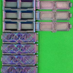 May include: A set of iridescent, multi-colored 3D printed pill organizers with hinged lids. The containers are labeled with the days of the week, specifically Wednesday, Thursday, Friday, and Saturday, featuring rectangular compartments for daily medication storage.