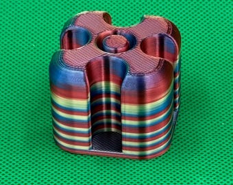 May include: A 3D printed square object with rounded corners and a central circular indentation. The piece features horizontal layers of gradient colors including red, blue, yellow, and green, creating a striped appearance throughout the structure.