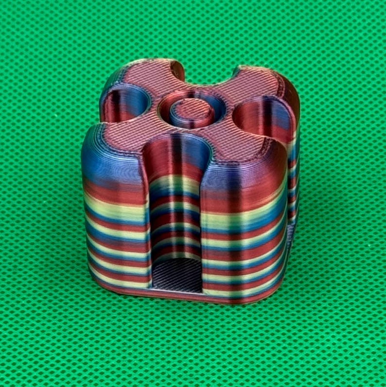 May include: A 3D printed square object with rounded corners and a central circular indentation. The piece features horizontal layers of gradient colors including red, blue, yellow, and green, creating a striped appearance throughout the structure.